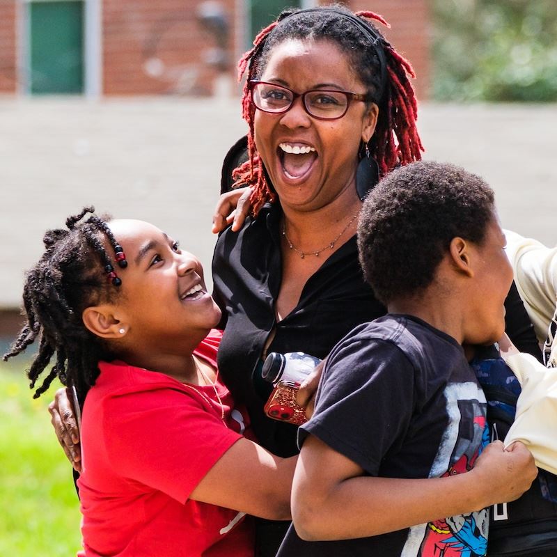  A teacher laughs while her students jump up and down around her.