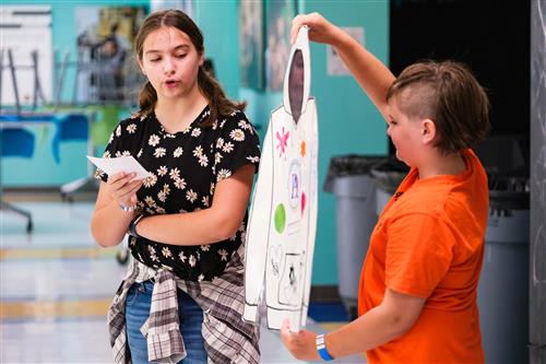 Girl and boy presenting a design project. Girl is standing facing the project boy is holding the design product