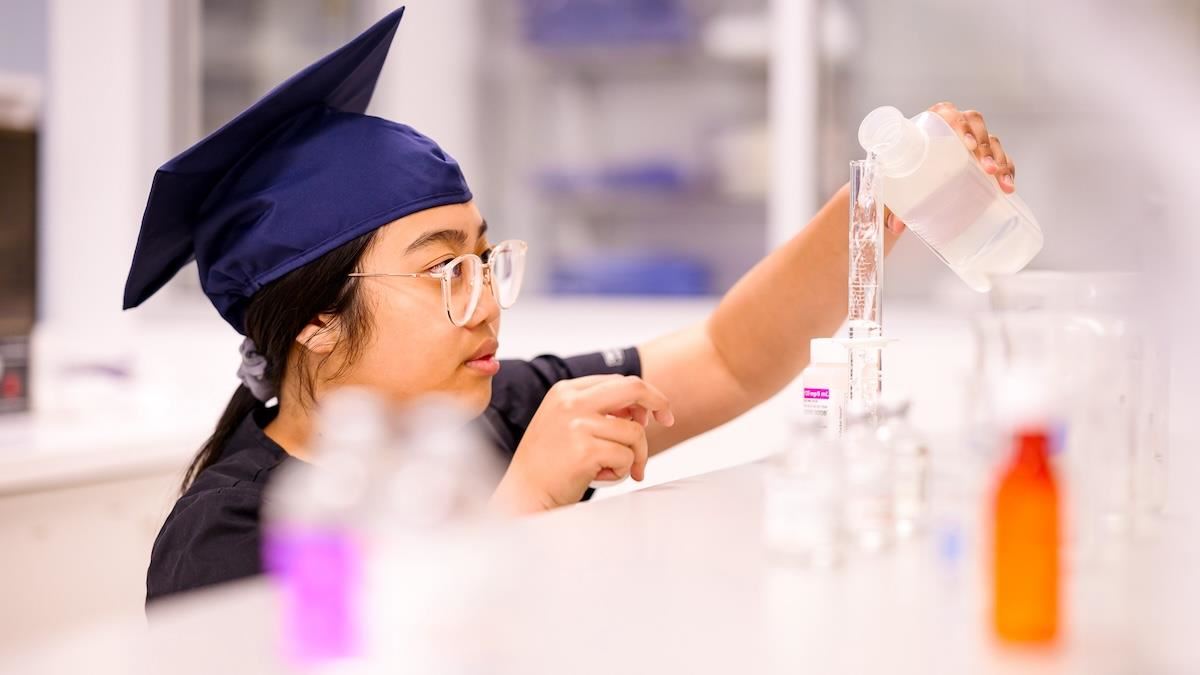 Phuong carefully pours a liquid into a glass cylinder